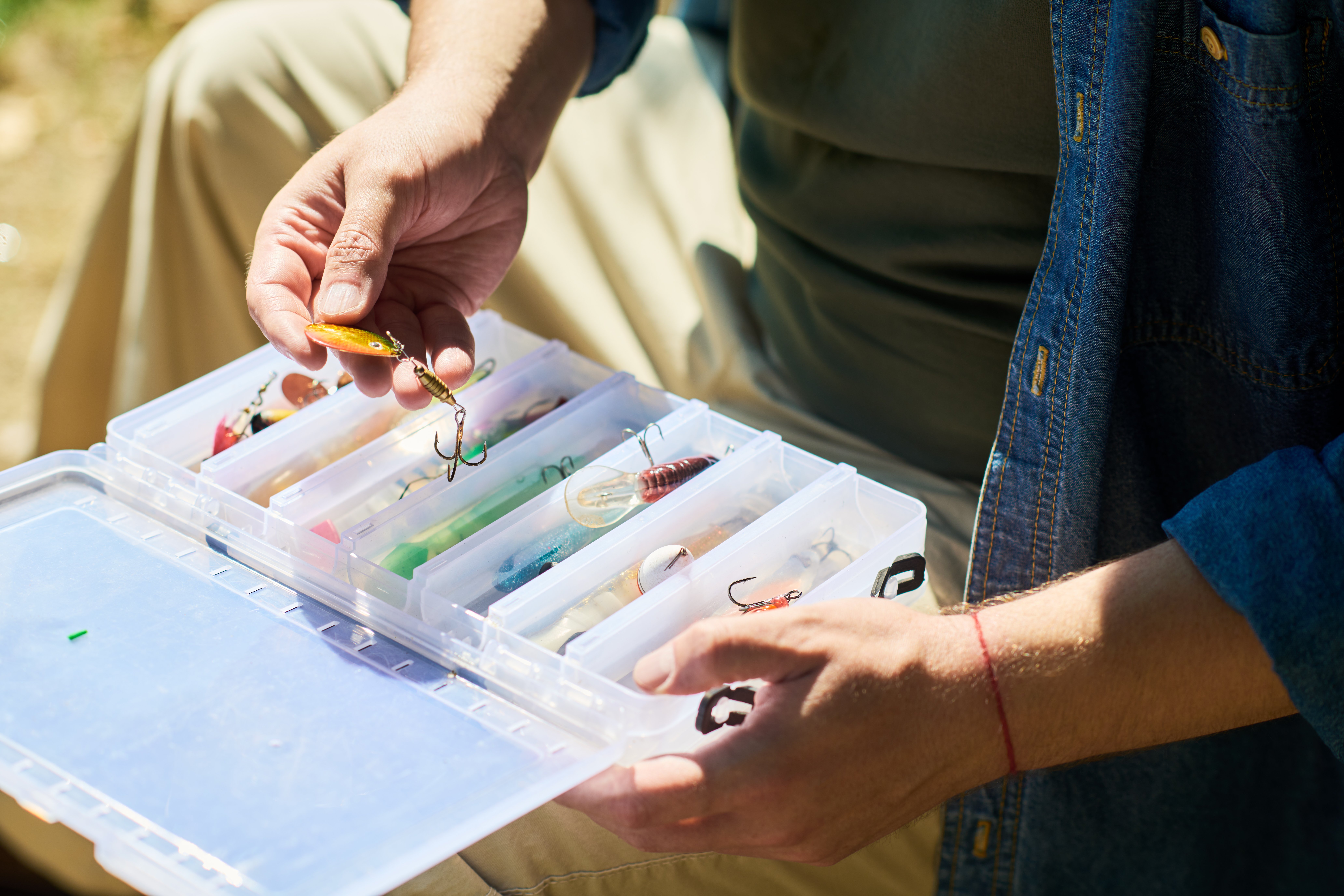 Close up of Caucasian young adult man holding lure over plastic tackle box outdoors. Outdoor recreation scene suitable for fishing gear marketing, hobby blogs, and sports catalogs
